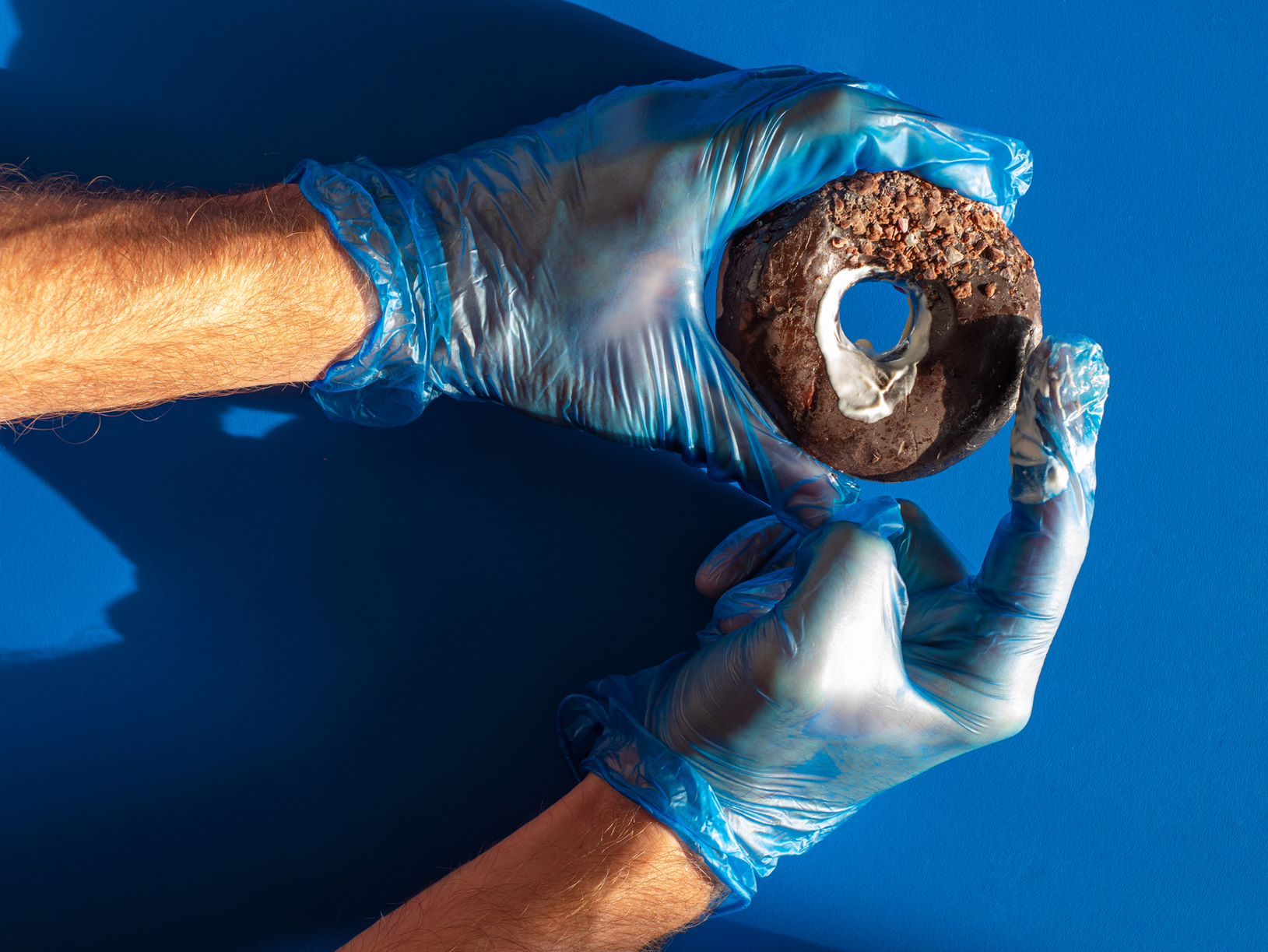 Männerhände mit blauen Handschuhen halten einen braunen Donut und schmieren diesen mit weißer Creme ein