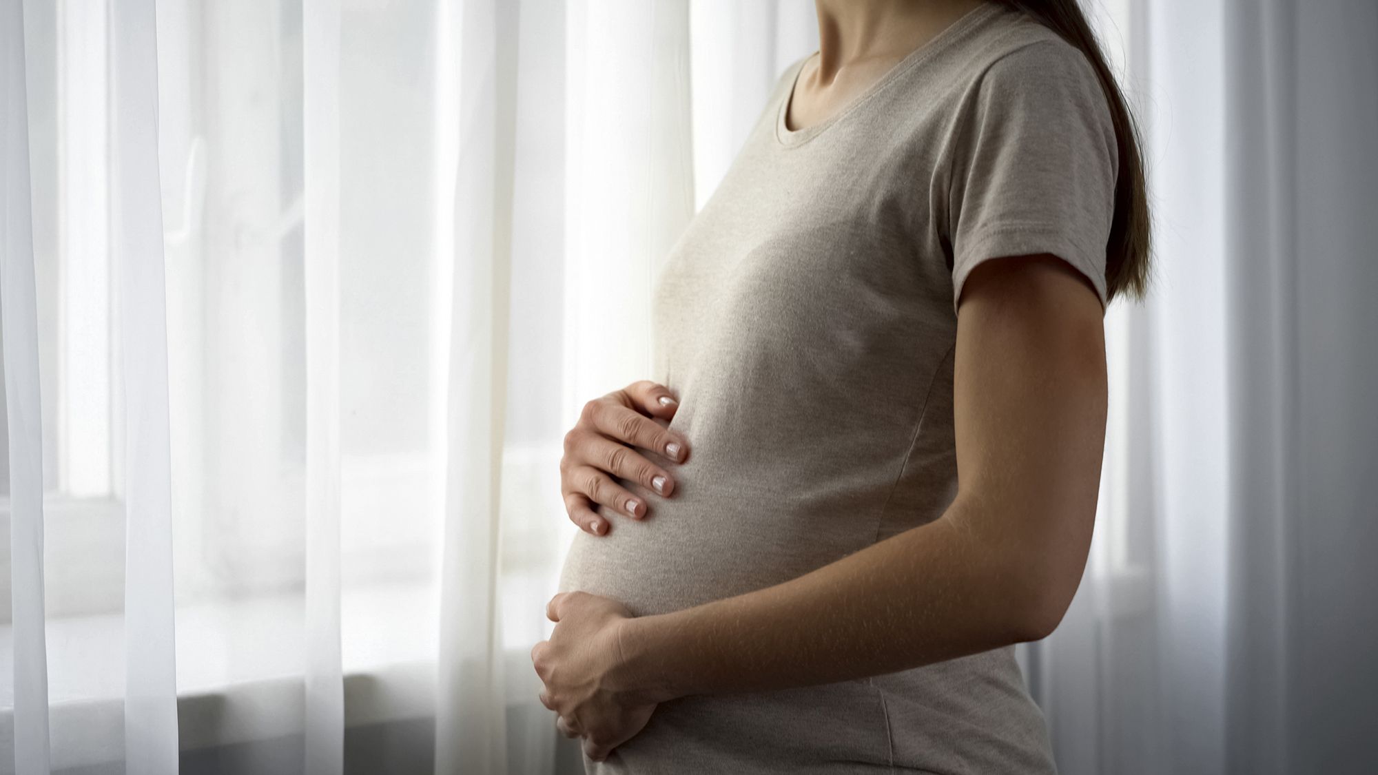 Schwangere Frau legt die Hände auf ihren Bauch und schaut aus dem Fenster