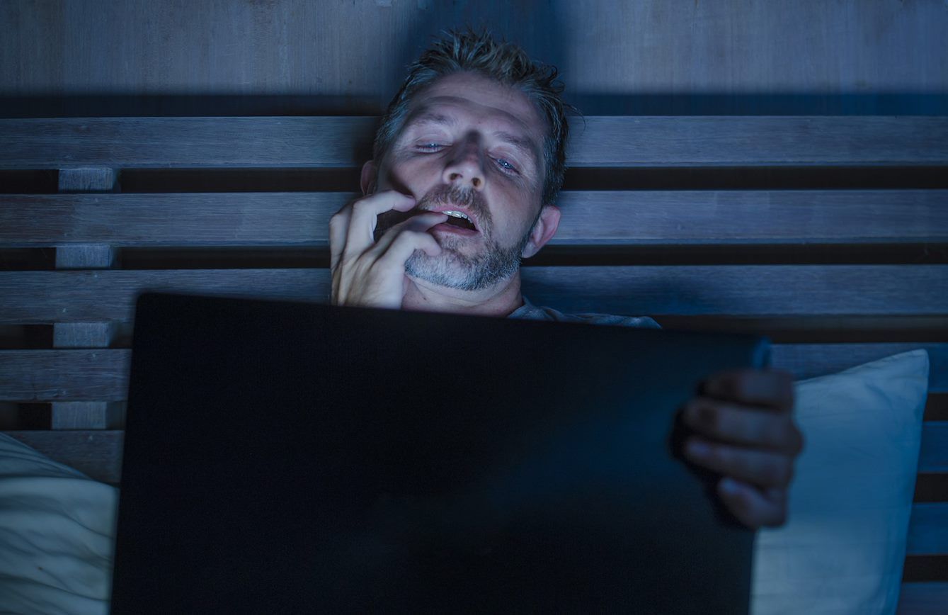 Mann sitzt im Bett, kaut an seinen Fingernägeln und schaut gespannt auf den Laptop vor sich..