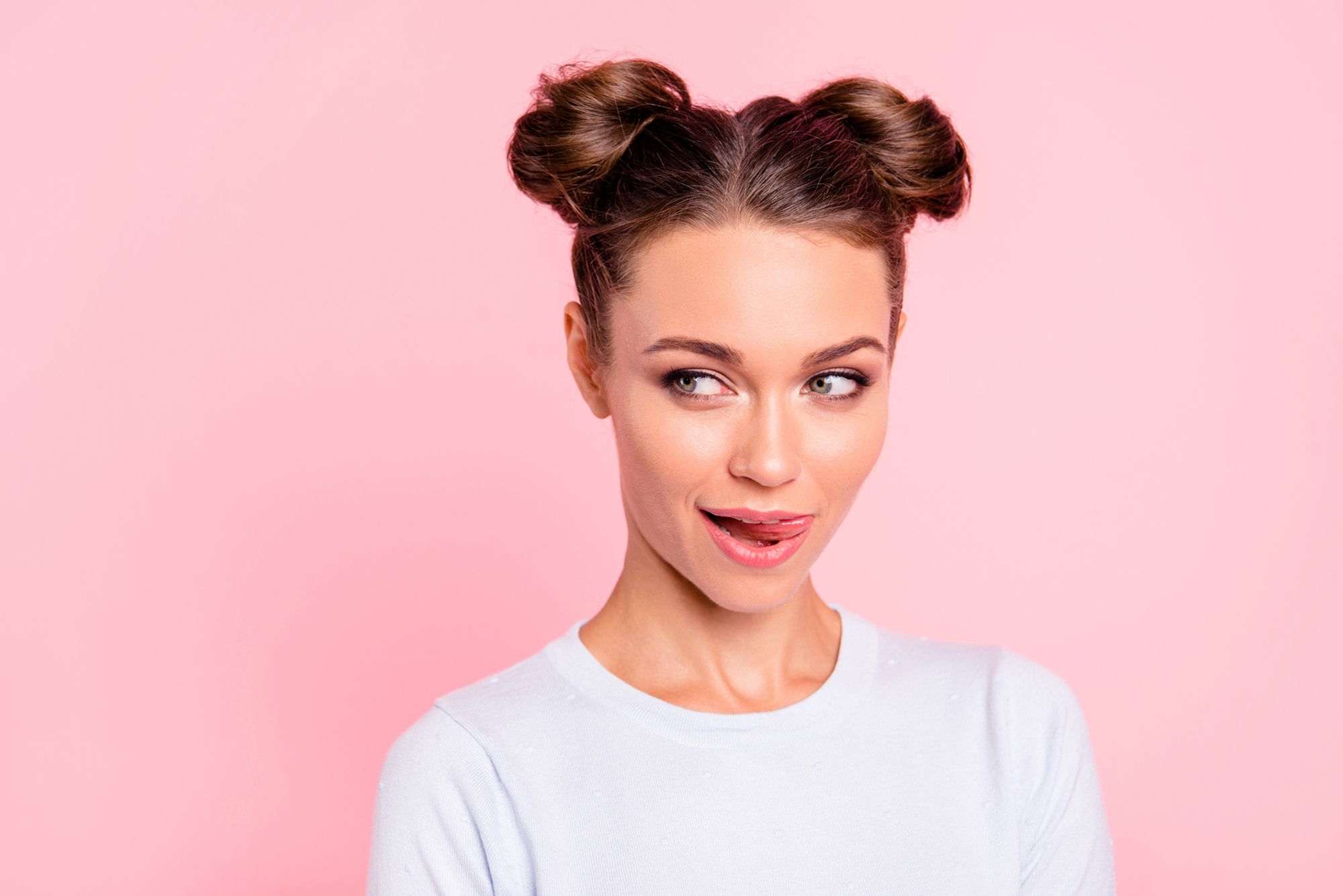 Brünette Frau in weißem Shirt und zwei Zöpfen, die sich über die Lippen leckt und zur Seite schaut vor rosa Hintergrund