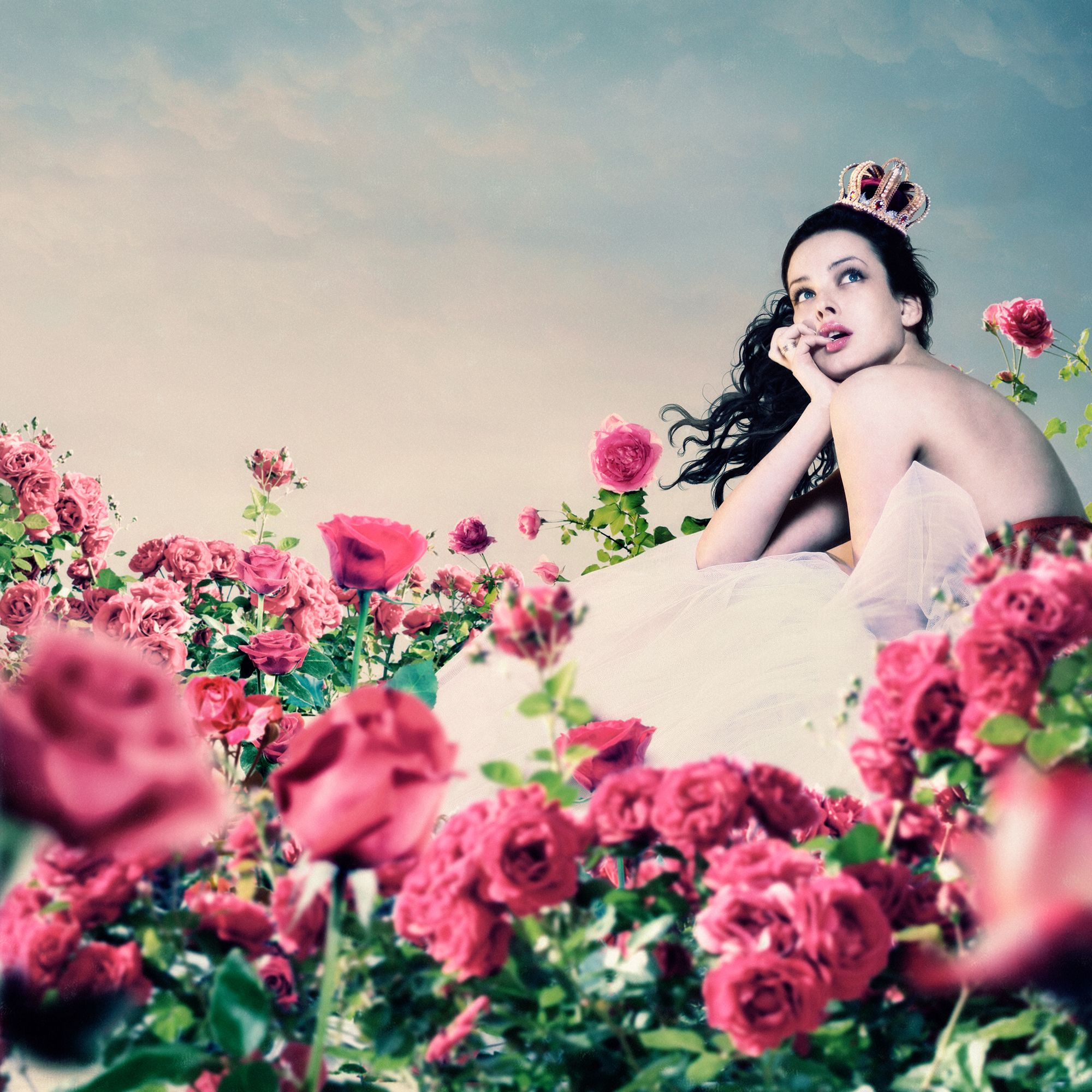 Schwarzhaarige Frau in weißem Kleid, die auf einer Wiese mit rosa Blumen sitzt und in den Himmel schaut