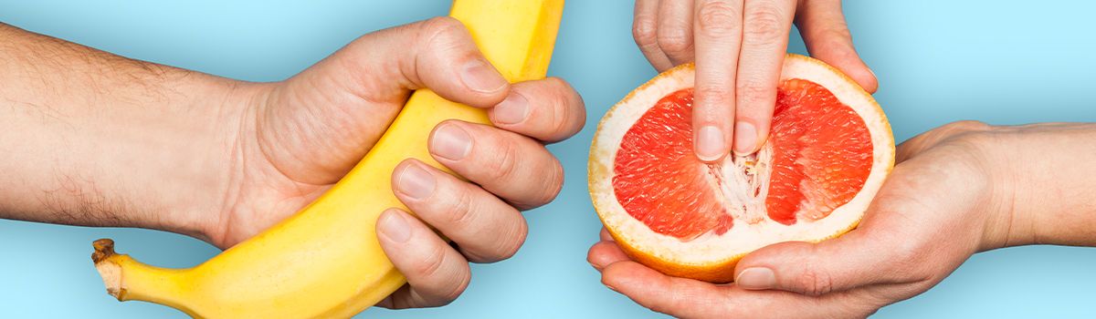 Mann hält eine Banane, Frau führt Finger in halbe Orange ein