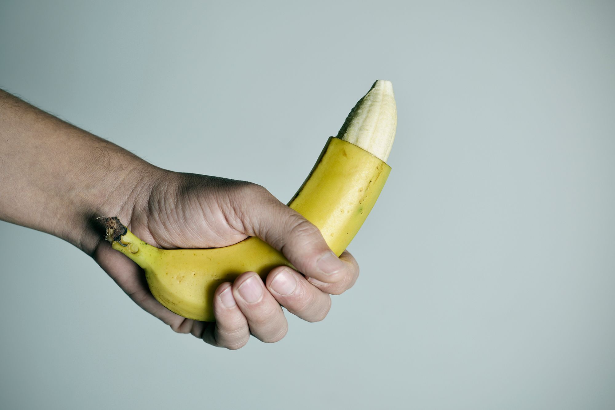 Hand mit einer Banane, bei der am oberen Ende die Schale abgeschnitten ist vor grauem Hintergrund