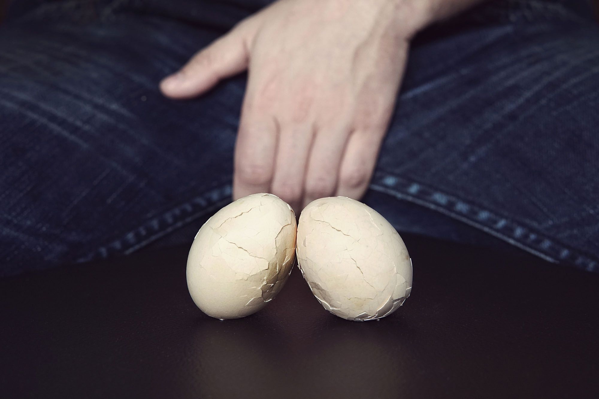 Zwei zerbrochene Eier mit einem Mann im Hintergrund, der seinen Schritt mit einer Hand hält