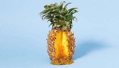 Eine teilweise aufgeschnittene Ananas aus der eine goldene Flüssigkeit herausläuft vor einem hellbla