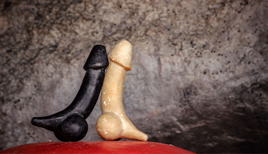 Ein schwarzer und ein naturfarbener Holzpenis die sich gegenüberstehen vor einem steinernen Hintergr