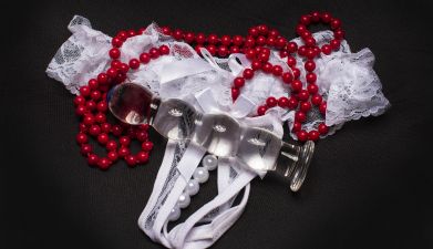 Ein weißer Spitzentanga, eine rote Perlenkette und ein Glasdildo vor einem schwarzen Hintergrund