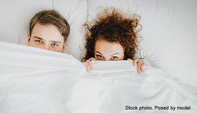 Ein Paar, das im Bett liegt, mit hochgezogener Decke, sodass nur noch die Augen rausschauen