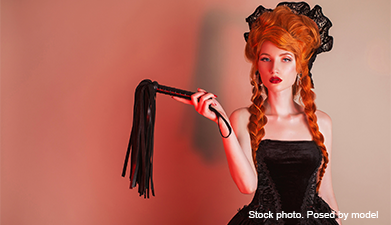 Eine Frau in Gothik-Kleidung mit einer roten Perücke auf und einer Peitsche in der Hand vor einem rö