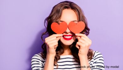Eine Frau vor einem lila Hintergrund die sich zwei rote Papierherzen vor die Augen hält