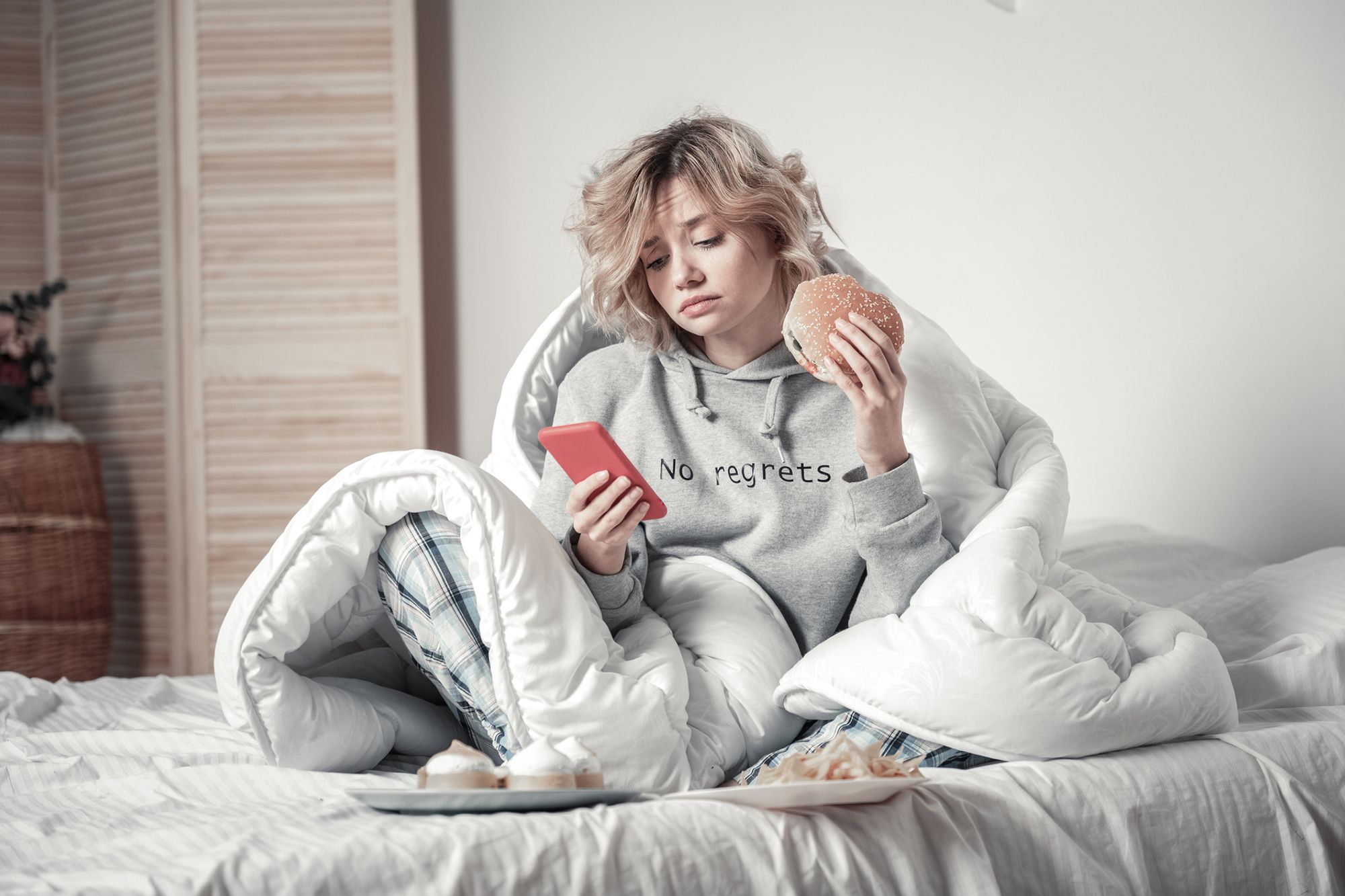 Eine Frau in einem grauen Hoodie, die in einer Decke gewickelt auf dem Bett sitzt, Fastfood isst und auf ihr Handy schaut