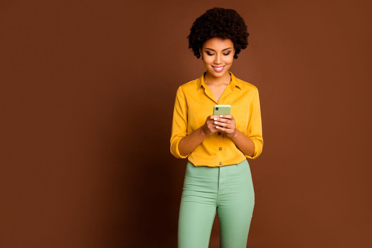 Frau, welche rücklings vor einer braunen Wand steht und lächelnd auf das Smartphone in ihren Händen schaut.