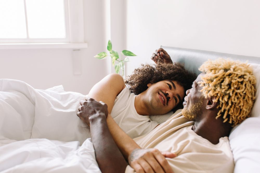 Zwei BIPoC Männer liegen Arm im Arm im Bett und sehen sich verliebt an
