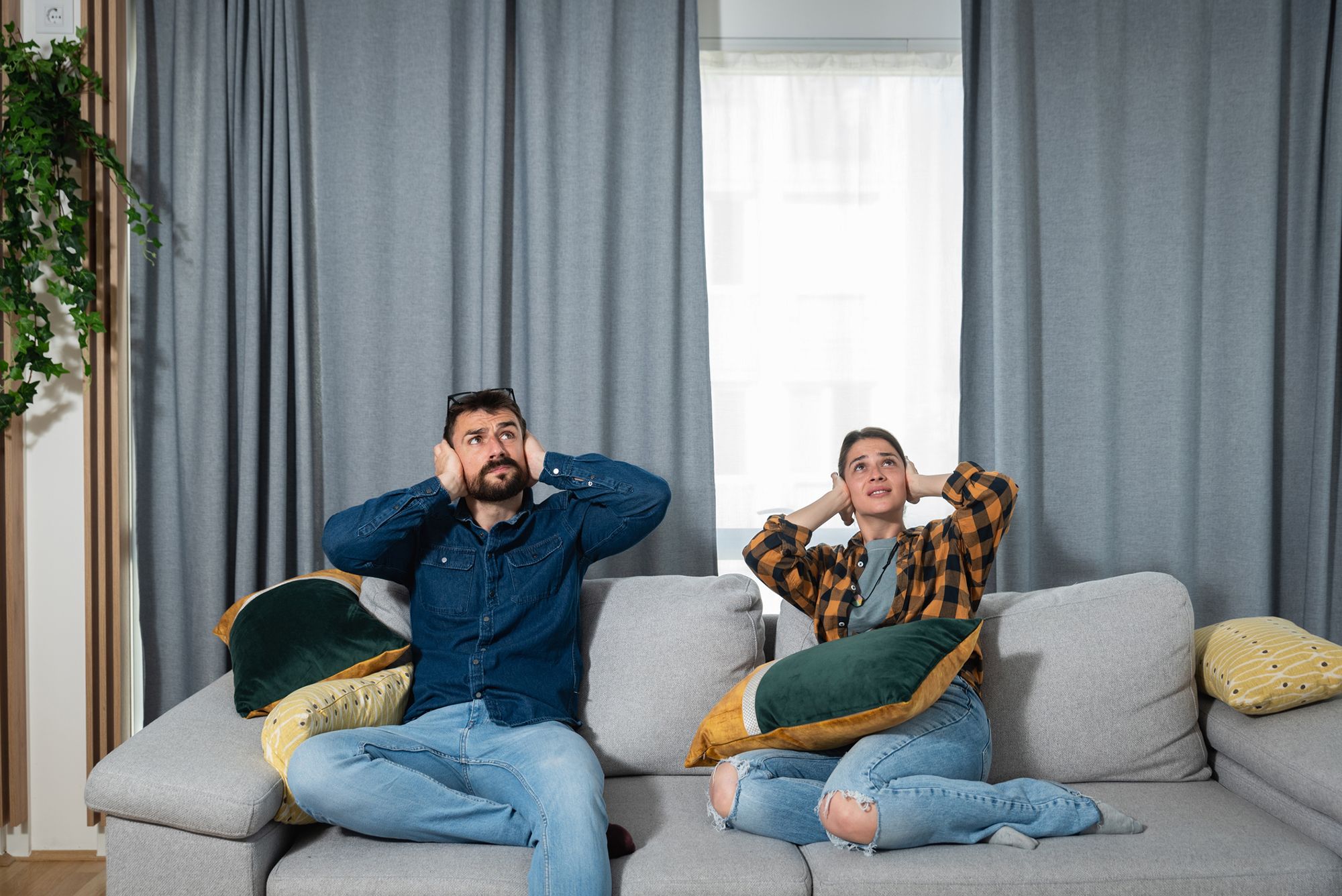 Ein Paar sitzt auf dem Sofa, blickt nach oben und hält sich die Ohren zu