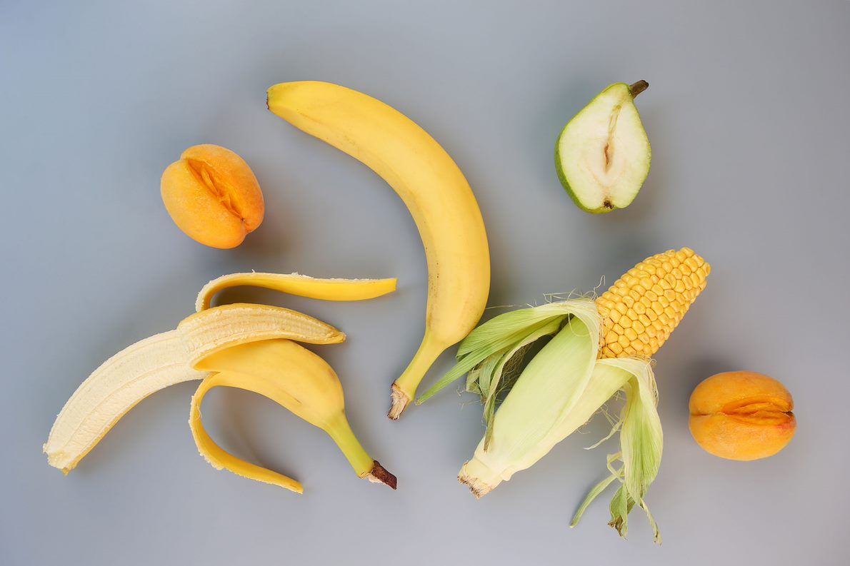 Zwei Bananen, Pfirsiche, ein Maiskolben und eine halbierte Birne verteilt auf grauem Hintergrund