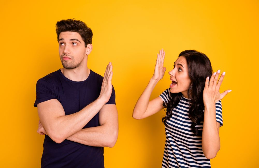Mann in schwarzem Shirt, der die Arme vor der Brust verschränkt, zur Seite schaut und einer Frau in gestreiftem Shirt die Hand vor das Gesicht hält vor gelbem Hintergrund