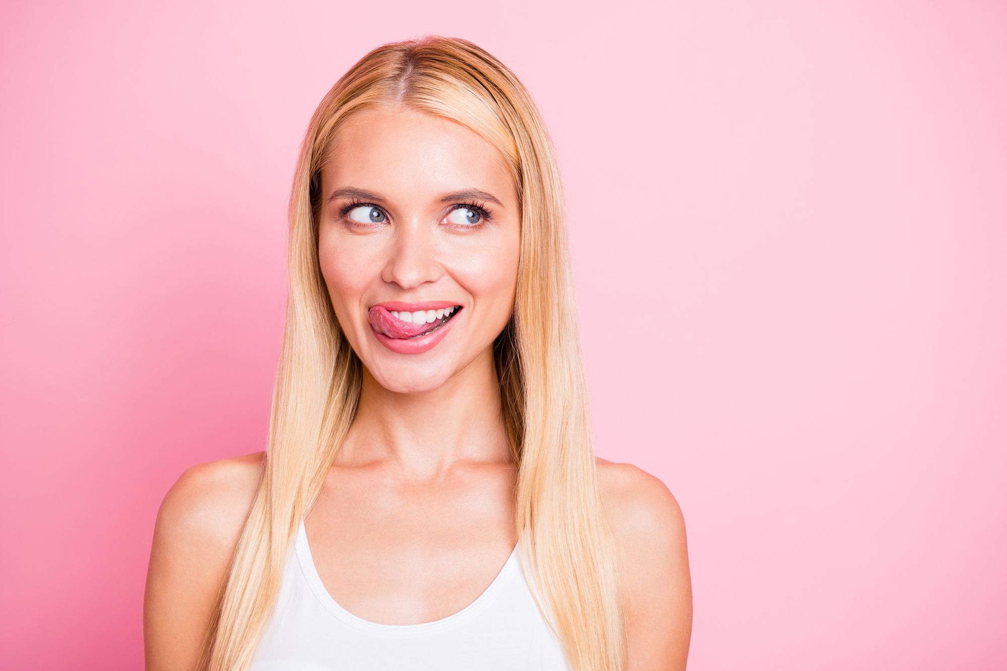 Eine blonde Frau die keck ihre Zunge herausstreckt vor einem rosafarbenen Hintergrund