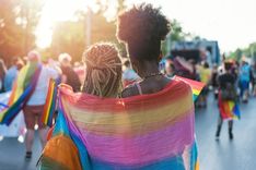 Zwei Frauen mit Regenbogenflagge auf einer Pride-Parade umarmen sich, aufgenommen beim Sonnenuntergang.