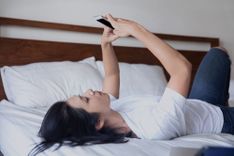 Frau liegt auf dem Rücken im Bett und hält das Handy in beiden Händen