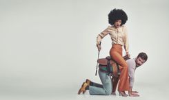 Frau in beiger Bluse, orangener Stoffhose und beigen High-Heels mit Reitgerte in der Hand, die auf dem Rücken eines Mannes in Hemd und Jeans reitet und ihm auf den Po haut