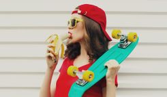 Brünette Frau in rotem Shirt und mit roter Snapback, die an einer Banane lutscht und ein Skateboard in der Hand hat vor einer weißen Häuserwand