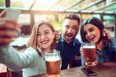 Brünette und schwarzhaarige Frau und brünetter Mann, die in einer Bar Bier trinken und ein Selfie schießen