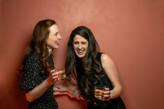 Zwei Frauen stehen mit Cocktailgläsern in der Hand nah beieinander, lachen und berühren sich am Arm