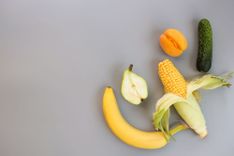 Banane, halbierte Birne, Pfirsich, Gewürzgurke und Maiskolben, die nebeneinander in der unteren rechten Ecke eines grauen Untergrunds liegen