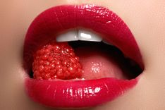 Frau mit roten Lippen hält eine Beere zwischen den Zähnen