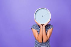 Eine Frau, deren Kopf durch eine Uhr ersetzt wurde und rücklings vor einer violetten Wand steht.