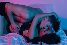 Frau in schwarzen Dessous liegt rücklings auf dem Bett und greift mit ihrer linken Hand in die Haare eines Mannes, welcher in Boxershorts über ihr kniet und sie küsst.