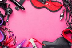 Schwarzer Knebel, schwarzer Dildo, rote Augenmaske, silberner Analplug mit rotem Stein, rote BDSM-Fesseln, schwarzes Paddle und lila Fesseln auf rosa Untergrund