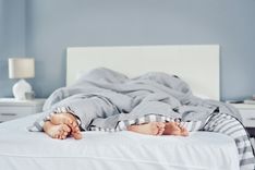 Zwei Menschen, die im Bett liegen und deren Füße unter einer grau-gestreiften Bettdecke und einer weiteren grauen Decke hervor schauen in einem Schlafzimmer