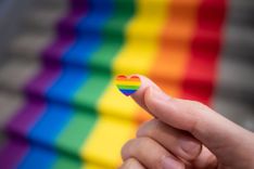 Herz mit Regenbogenmuster liegt auf einem Zeigefinger, Regenbogenfahne im Hintergrund