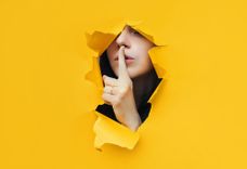 Frau hält den Zeigefinger vor dem Mund und ist hinter einem gelben Plakat versteckt
