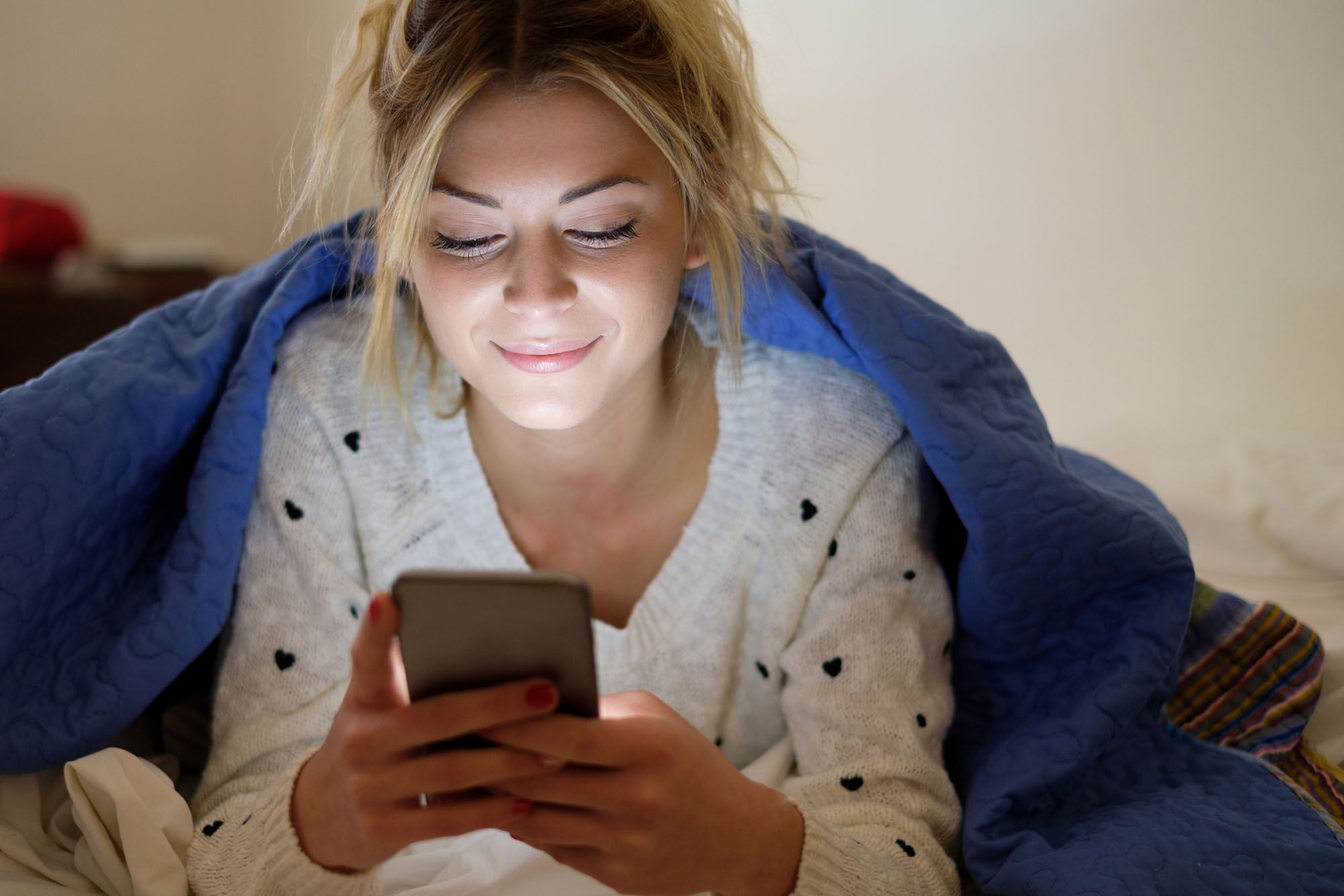 Frau in grauem Pullover mit schwarzen, kleinen Herzen und blauer Decke ummantelt, schaut auf ihr Handy, dass sie in den Händen hält