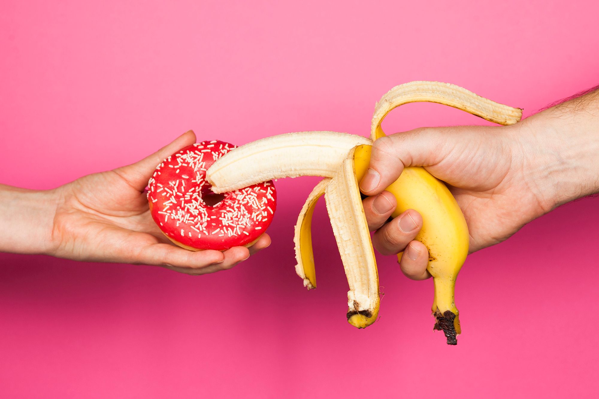 Eine Hand, die eine halb-geschälte Banane über einen Donut hält, der von einer anderen Hand gehalten wird auf pinkem Hintergrund